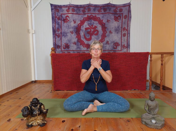 Vrouw zit in kleermakerszit op een yogamat en houdt haar handen tegen haar keel in een meditatie- of ademhalingsoefening.