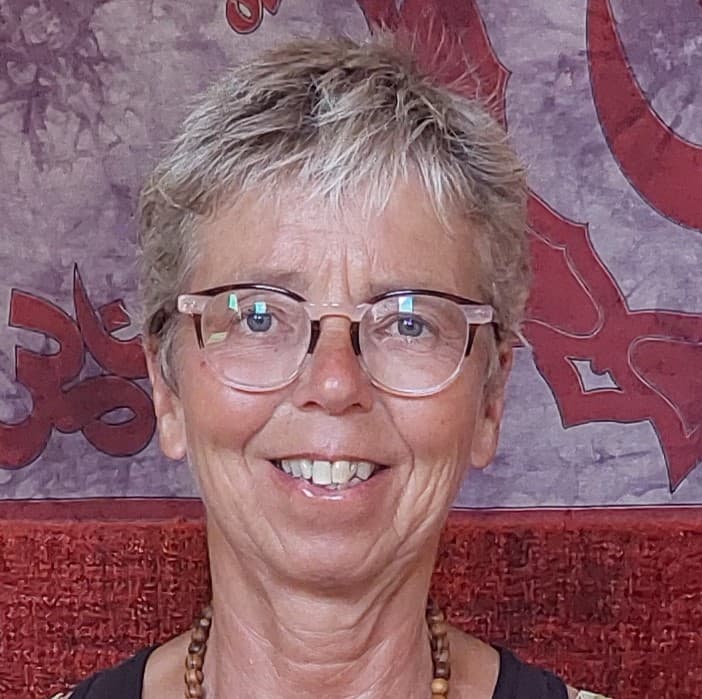 Vrouw met grijs haar en bril glimlacht voor een kleurrijk achtergrondtapijt met rode patronen.