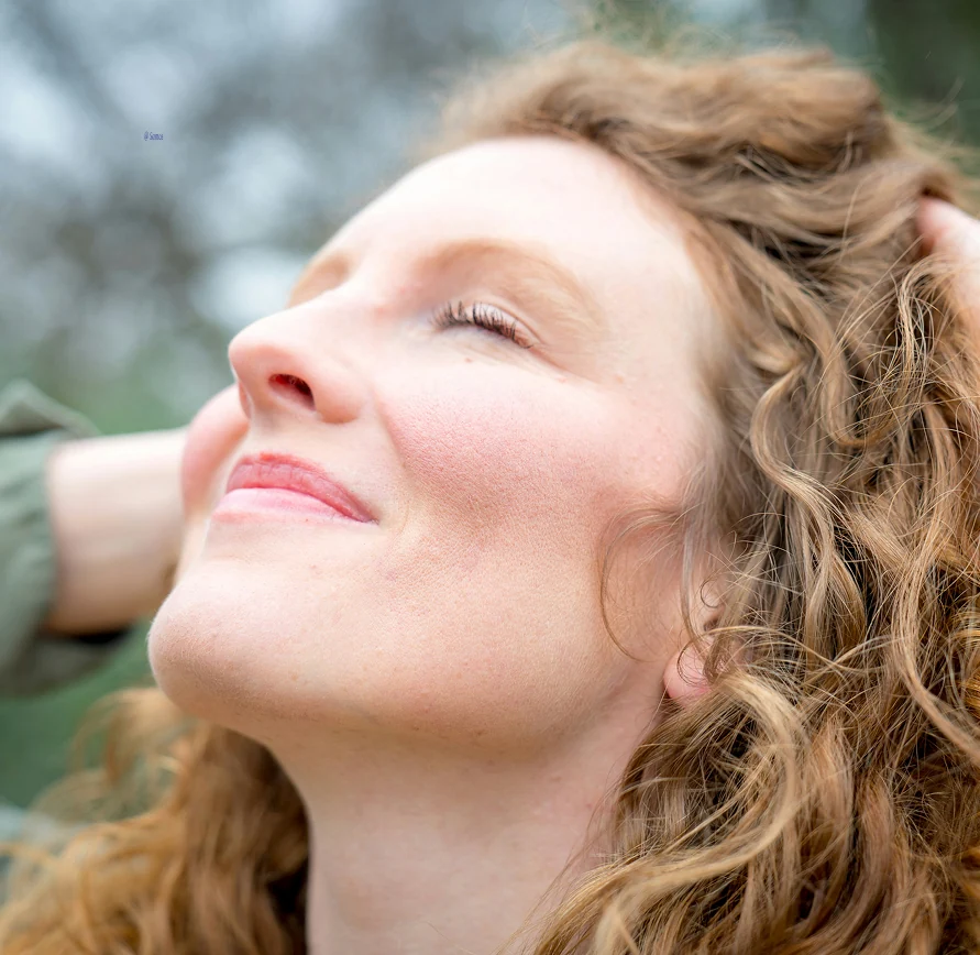 Vrouw met krullend haar sluit ontspannen haar ogen en glimlacht zacht in een moment van rust.