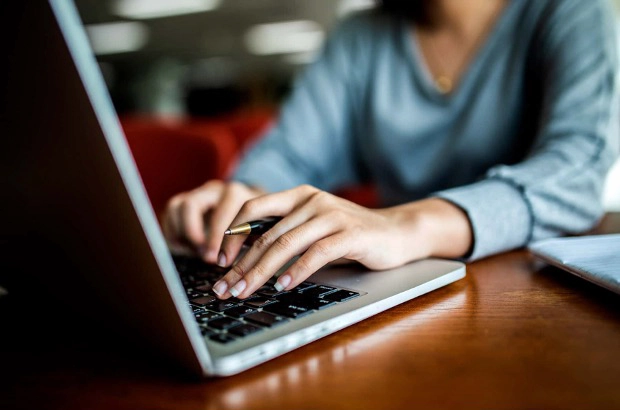 Een persoon typt op een laptop terwijl die een pen vasthoudt, zittend aan een houten tafel.