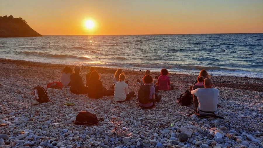 Groep mensen zit op een kiezelstrand tijdens zonsondergang bij de zee, onderdeel van Marjaani.