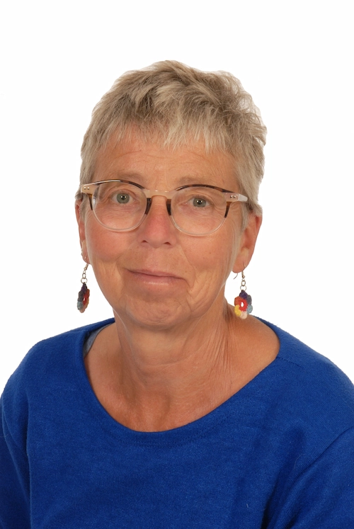 Portretfoto van een vrouw met kort grijs haar, een blauwe trui en bloemvormige oorbellen tegen een witte achtergrond.