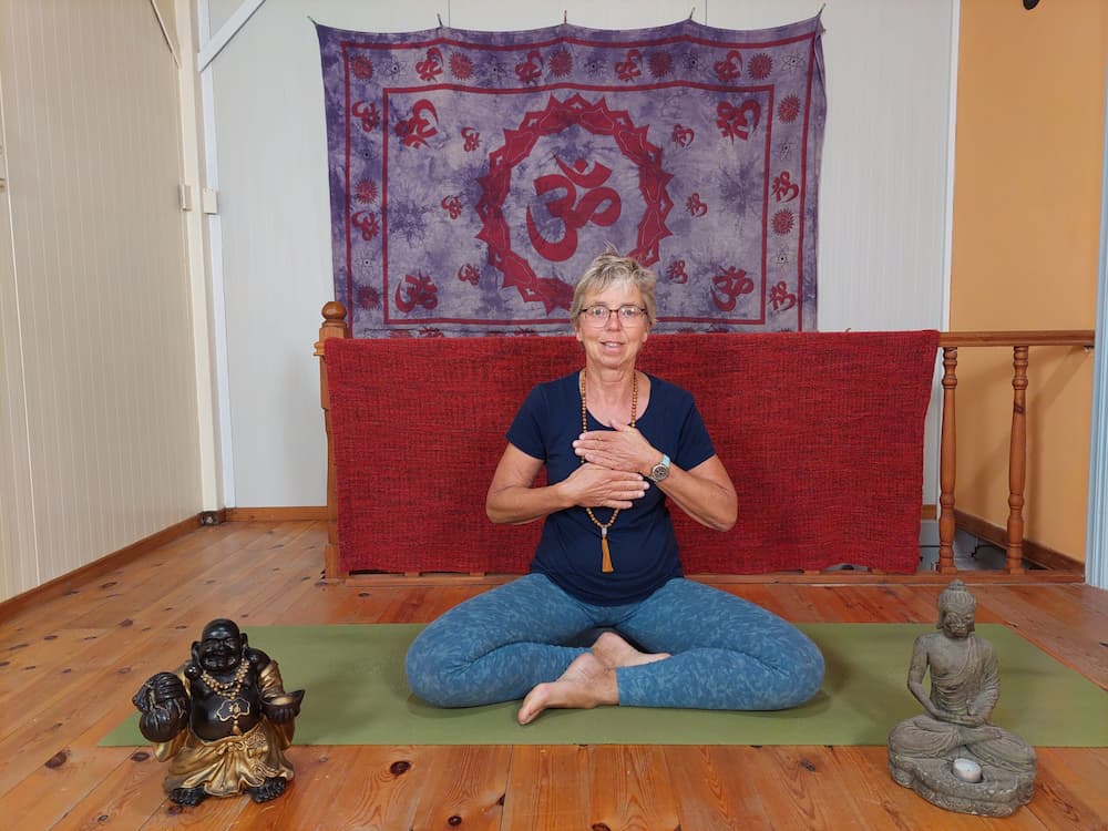 Vrouw in kleermakerszit mediteert met handen op het hart op een yogamat voor een doek met het symbool van het hartchakra.