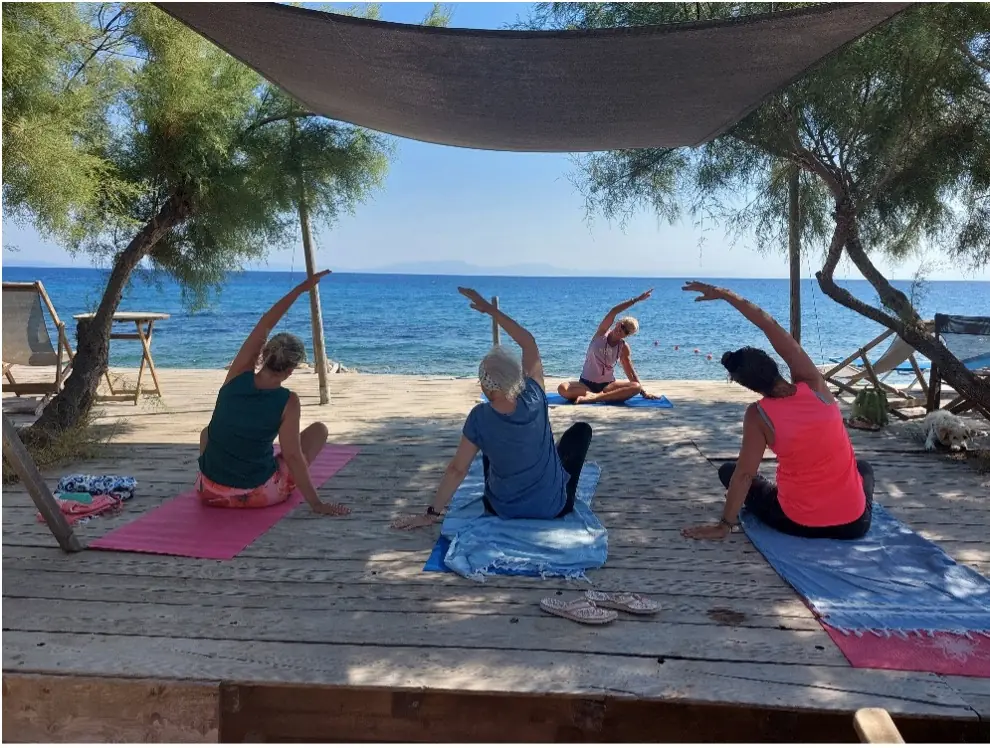 Vier vrouwen volgen een yogales van Marjaani Yoga op een houten vlonder aan zee onder schaduwdoeken.