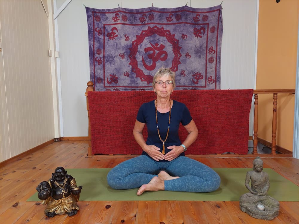 Vrouw zit in kleermakerszit op een yogamat en focust op het sacraalchakra met handen op de onderbuik.