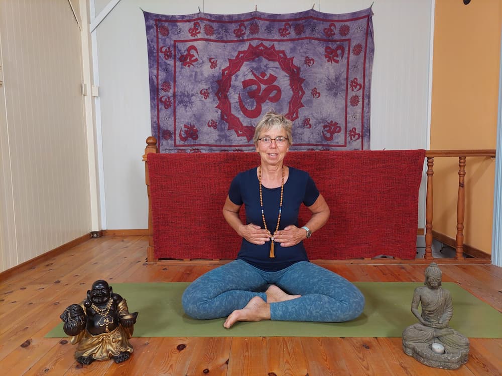 Vrouw in kleermakerszit op een yogamat wijst naar haar zonnevlecht tijdens een yogasessie, met boeddhabeelden en een paarse wanddoek op de achtergrond.