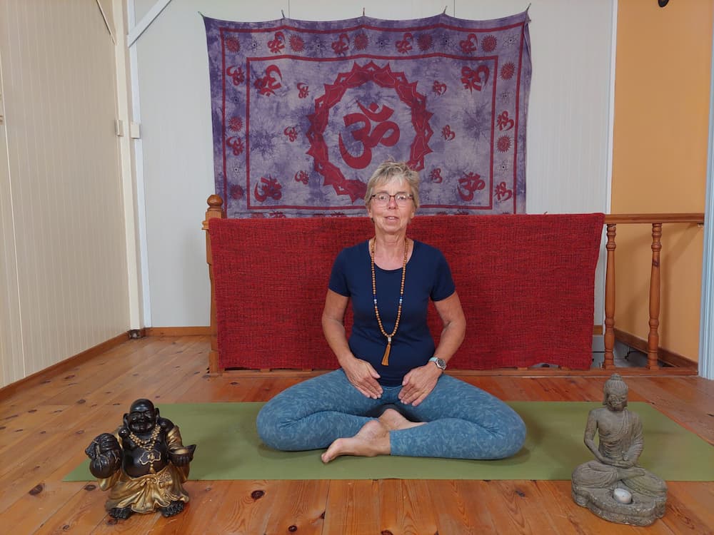 Vrouw in lotushouding mediteert op een yogamat voor een wandkleed met symbool van het wortelchakra.